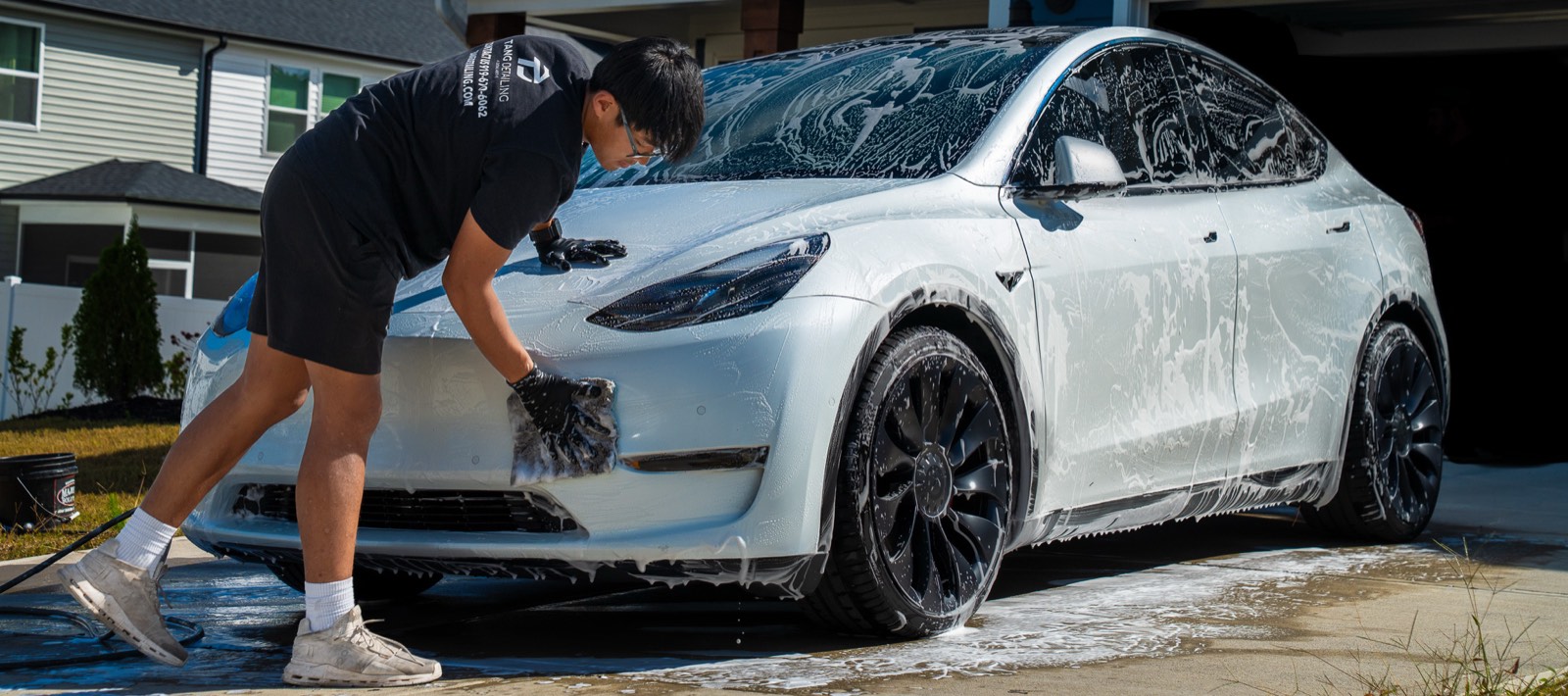 Professional detailer hand-washing a foam-covered white Tesla Model Y in a residential driveway