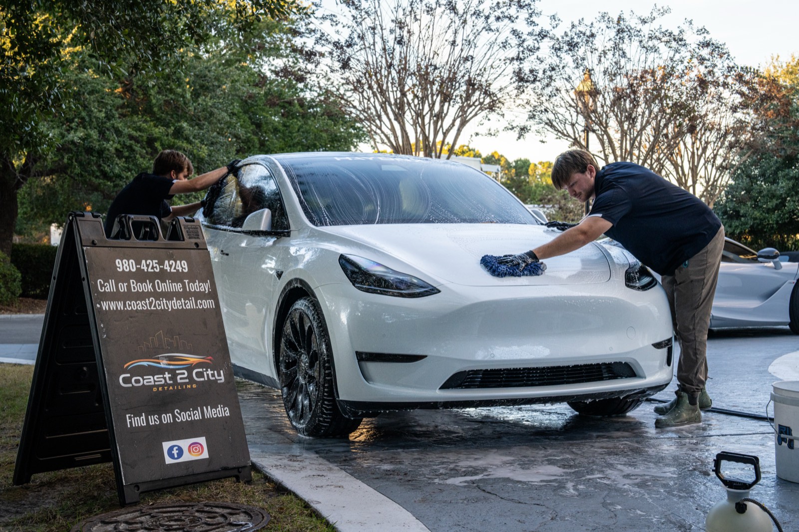 Two detailers hand-washing a white Tesla Model Y beside a detailing shop sign