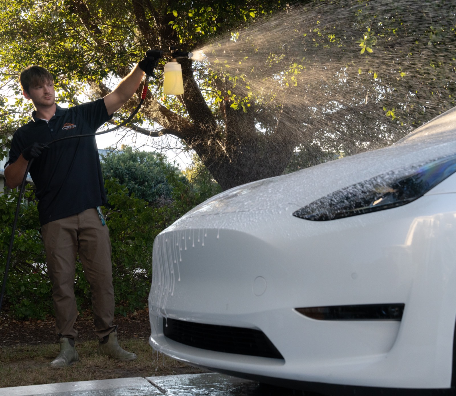 Detailer applying snow foam over a white Tesla front end with sun-backlit spray at golden hour