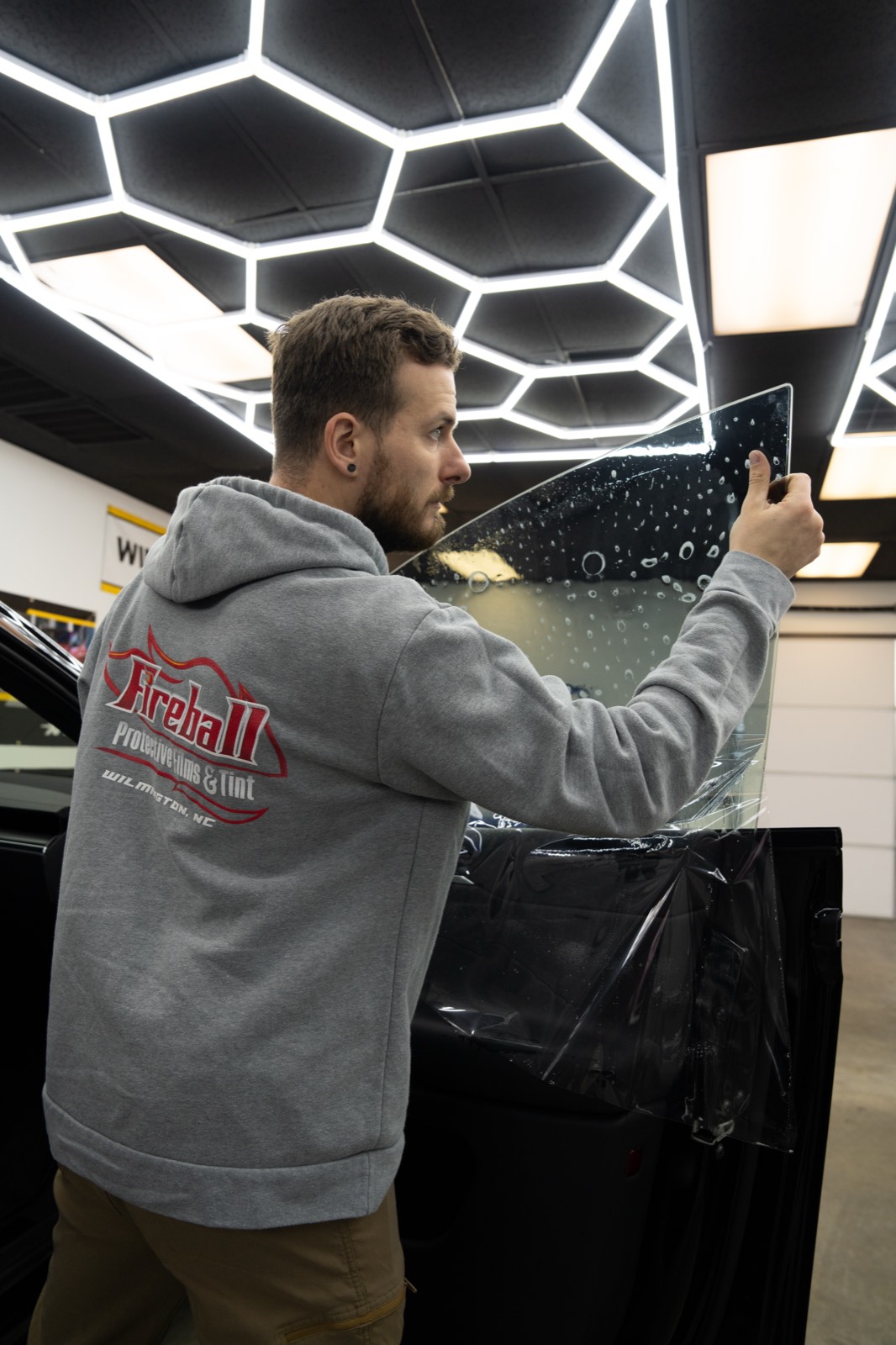 Technician handling a sheet of window tint film during installation in a hexagon-lit studio